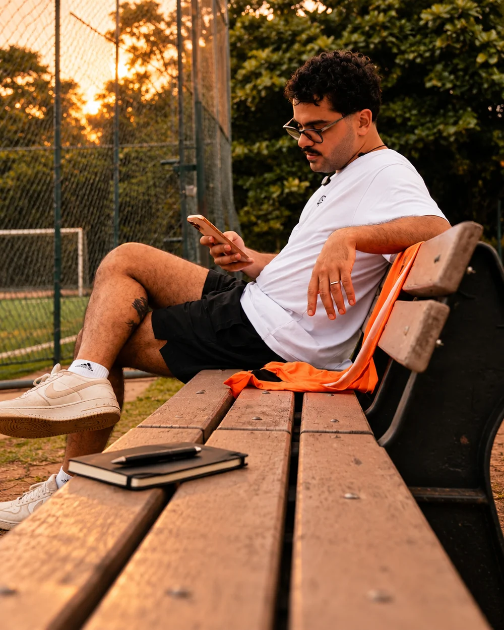 Organizador consulta o celular no banco ao lado do campo, com um caderno de anotações por perto